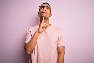 Yakışıklı Afro-Amerikalı adam pembe arka planda günlük tişört ve gözlük takıyor. Kuşkuya odaklanmış, çenesinde bir parmak ve yukarı bakıp merak ediyor.