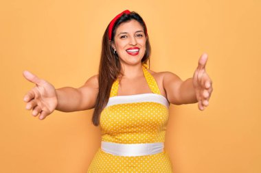 50 'li yaşlarda modaya uygun seksi giyinen genç Latin bir kadın sarı arka planda kameraya sarılıp gülümsüyor. Mutluluğu kucaklayan neşeli bir ifade..