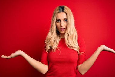 İzole edilmiş kırmızı arka planda duran ve kolları açık duran gündelik tişört giyen genç ve güzel sarışın bir kadın..