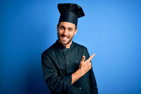 年轻英俊的厨师 留着胡子 身穿炊具制服 头戴蓝色背景的帽子 面带微笑 手指手画脚地朝旁边看去 脸上带着愉快而自然的表情 — 图库照片