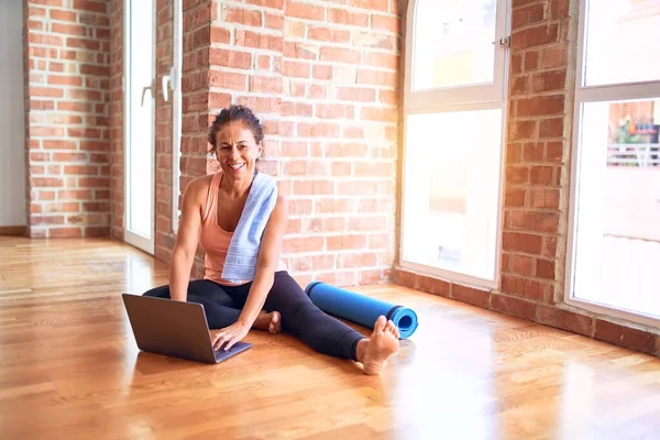 Middle age beautiful sportwoman smiling happy and confident. Sitting on the floor with smile on face using laptop before doing exercise at gym