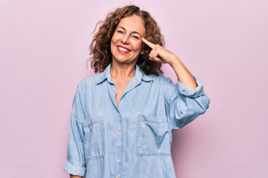 Orta yaşlı güzel bir kadın pembe arka planda sıradan bir kot gömlek giyiyor. Tek parmağıyla gülümsüyor, harika bir fikir ya da düşünce, iyi bir hafıza.