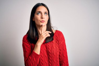 Mavi gözlü genç esmer kadın izole edilmiş beyaz arka plan üzerine gündelik süveter giyiyor. Şüphe üzerine yoğunlaşmış, çenesi parmaklarla yukarı bakıyor ve merak ediyor.