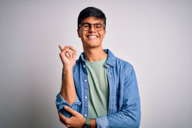 İzole edilmiş beyaz arkaplanda gözlüklü ve sıradan bir gömlek giyen genç yakışıklı bir adam. Yüzünde kocaman bir gülümseme, eli ve parmağı kameraya bakıyor..