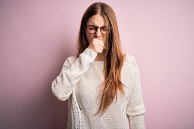 Genç, güzel, kızıl saçlı bir kadın pembe arka planda günlük süveter ve gözlük takıyor kendini iyi hissetmiyor ve soğuk algınlığı veya bronşitin belirtisi olarak öksürüyor. Sağlık hizmeti kavramı.