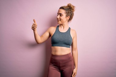 Genç ve güzel sarışın sporcu kadın spor yapıyor. Pembe arka planda spor giysisi giyiyor. Gururlu görünüyor, gülümsüyor.