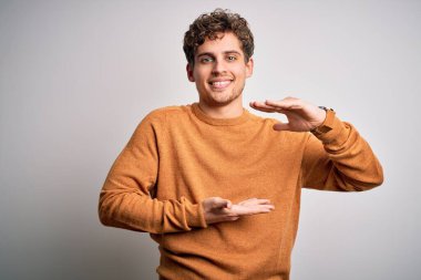 Sarışın, kıvırcık saçlı, beyaz arka plan üzerine gündelik süveter giyen, elleri büyük ve büyük boy işareti gösteren, ölçü sembolü olan genç, yakışıklı bir adam. Kameraya bakıp gülümsüyor. Ölçüm konsepti.