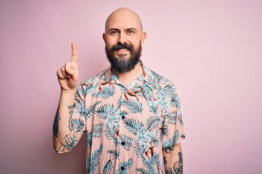 Sakallı, dövmeli, pembe arka planda sıradan bir çiçek gömleği giyen yakışıklı kel adam başarılı bir fikri işaret ediyor. Heyecanlı ve mutlu. Bir numara..