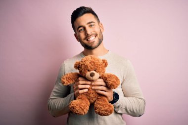 Genç, yakışıklı bir adam, ayıcığı pembe arka planda tek başına dikiliyor. Mutlu bir yüzü var, gülümsüyor ve kendine güvenen bir gülümsemeyle dişlerini gösteriyor.