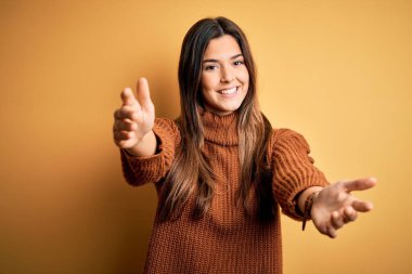 İzole edilmiş sarı arka planda dikilip sarılmak için kollarını açmış gülümseyen genç ve güzel bir kız. Mutluluğu kucaklayan neşeli bir ifade..