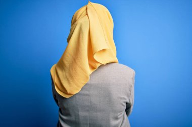 Orta yaşlı esmer iş kadını, geleneksel Müslüman tesettüre bürünmüş mavi arka planda geriye doğru bakarak çapraz kollara bakıyor.