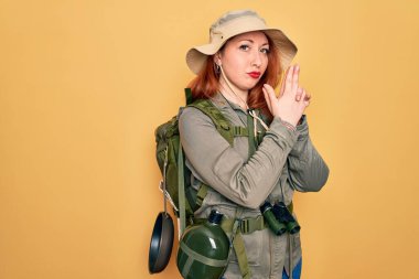 Sırt çantalı, sarı şapkalı, sırt çantalı genç kızıl saçlı kadın. Sembolik bir silah tutuyor. El hareketi yapıyor.