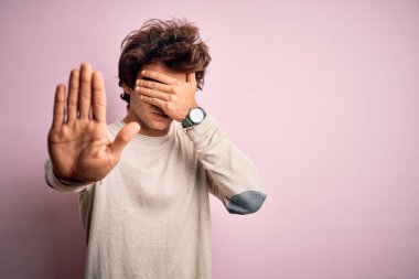 Genç ve yakışıklı bir adam, günlük tişört giyip pembe arka planda dikiliyor, gözleri ellerle kaplıyor ve üzgün ve korku ifadeleriyle jest yapıyor. Utanç verici ve olumsuz bir kavram..