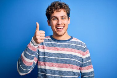 Sarışın, kıvırcık saçlı, mavi arka planda çizgili süveter giyen ve el hareketleriyle mutlu mesut hareket eden genç bir adam. Kameraya bakıp başarıyı gösteren ifade onaylanıyor.