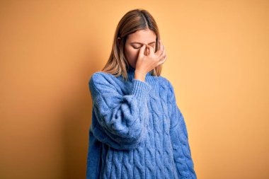 Sarıya boyanmış arka plan üzerine balıkçı yaka kazak giyen genç, güzel sarışın kadın yorgun burnu ve gözleri ovuşturarak yorgunluk ve baş ağrısı hissediyor. Stres ve hüsran kavramı.