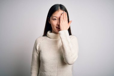 Genç, güzel Asyalı bir kadın, günlük süveter giyiyor, izole bir arka planda duruyor, bir gözünü eliyle kapatıyor, yüzünde kendine güvenen bir gülümseme ve sürpriz bir duygu var..