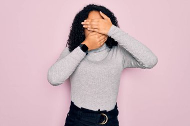 Genç Afro-Amerikalı kadın balıkçı yaka kazak ve pembe arka plan üzerine gözlük takıyor gözleri ve ağzı kapalı, şaşırmış ve şok olmuş. Duygularını gizliyor.