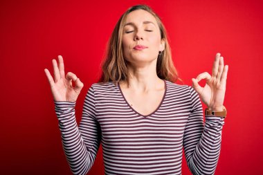 İzole edilmiş kırmızı arka plan üzerine sıradan çizgili tişört giyen genç ve güzel sarışın kadın rahatla ve gözün kapalı gülümse parmaklarınla meditasyon hareketi yap. Yoga konsepti.
