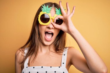 Sarı arka planda ananas güneş gözlüğü takan genç, güzel esmer kadın gülümsüyor.