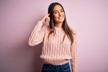 Genç, güzel esmer kadın, pembe arka planda günlük süveter giyiyor. Tek parmakla kafasını göstererek gülümsüyor. Harika bir fikir ya da düşünce.