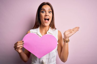 Genç, güzel, esmer bir kadın elinde kocaman pembe bir kalp kâğıdıyla sevgililer gününü kutluyor. Çok mutlu ve heyecanlı. Kazanan ifade zaferi kutluyor.