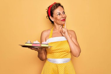 Orta yaşlı, son sınıf rozetli, 50 'lerin stili retro elbise giyen, elinde kap kek tepsisi olan, ciddi yüzlü soru soran, kafası karışık bir kadın.