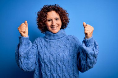 Orta yaşlı güzel kıvırcık saçlı kadın mavi arka plan üzerine balıkçı yaka kazak giyip kollarını kaldırarak ve açık gözlerle başarıyı kutluyor. Kazanan konsept.