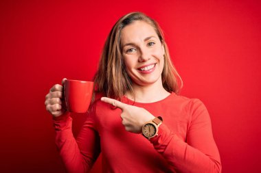 Genç, güzel, sarışın, kahve içen, izole edilmiş kırmızı arka planda duran, el ve parmağıyla işaret ederek mutlu bir şekilde duran bir kadın.