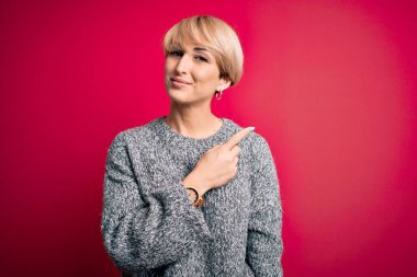 Modern kısa saçlı, pembe arka planda sıradan bir süveter giyen genç, sarışın bir kadın. El ile işaret ederek reklamını yapıyor. Ciddi ve sakin bir yüz ifadesi var.