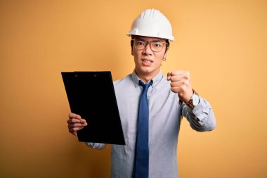 Güvenlik kaskı takan genç, yakışıklı Çinli mühendis elinde pano tutarken sinirlenmiş ve sinirlenmiş bir şekilde bağırıyor.