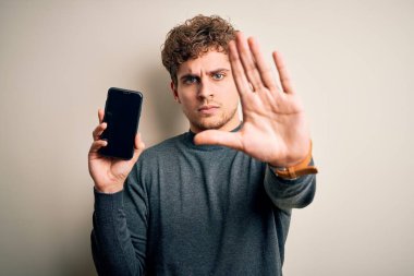 Sarışın, kıvırcık saçlı, akıllı telefon gösteren genç bir adam izole edilmiş beyaz arka planda duruyor. Eli açık, dur işareti yapıyor. Ciddi ve kendinden emin bir ifadesi var.