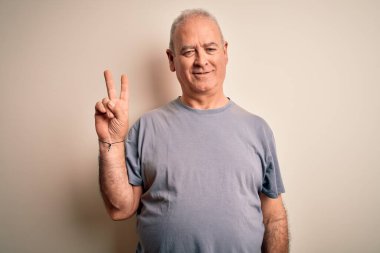Orta yaşlı, beyaz tişörtlü yakışıklı adam izole edilmiş beyaz arka planda dikiliyor. Kameraya gülümsüyor ve zafer işareti yapıyor. İki numara..