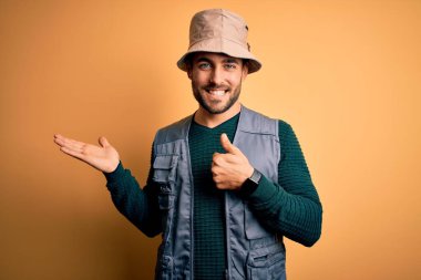 Tatilde sakallı yakışıklı bir turist sarı arka planda kaşif şapkası takıyor, el hareketi yapıyor, mutlu ve neşeli gülümsüyor.