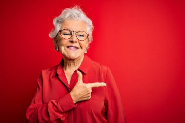Son sınıf güzeli, gri saçlı, rahat gömlekli, kırmızı arka planda gözlüklü neşeli yüzlü, yüzünde mutlu ve doğal bir ifade olan bir gülümseme olan bir kadın.