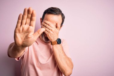 Orta yaşlı, beyaz tişörtlü bir adam izole edilmiş pembe arka planda dikiliyor gözleri ellerle örtüyor ve üzgün ve korku ifadeleriyle jest yapıyor. Utanç verici ve olumsuz bir kavram..