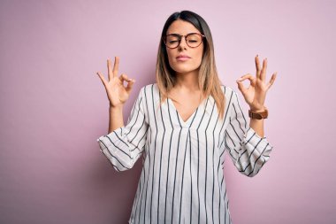 Genç ve güzel bir kadın, pembe arka plan üzerine günlük çizgili tişört ve gözlük takıyor rahatlamış ve gözleri kapalı gülümsüyor parmaklarıyla meditasyon hareketi yapıyor. Yoga konsepti.