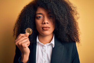Afro saçlı, Bitcoin 'i sarı arka planda tutan Afro-Amerikalı genç bir iş kadını akıllı yüzünde kendine güvenen bir ifadeyle ciddi düşünüyor.