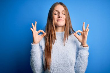 Genç, güzel, kızıl saçlı bir kadın izole edilmiş mavi arka plan üzerine günlük süveter giyiyor rahatlamış ve parmaklarıyla meditasyon hareketi yaparken gözleri kapalı gülümsüyor. Yoga konsepti.