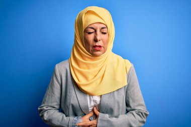 Orta yaşlı esmer iş kadını, geleneksel Müslüman tesettüre bürünmüş, mavi arka planda, midesi üstünde, midesi ağrıyan, hazımsızlık çeken, ağrılı bir hastalık yüzünden. Ache kavramı.