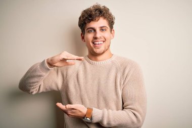 Sarışın, kıvırcık saçlı, beyaz arka plan üzerine gündelik süveter giyen, elleri büyük ve büyük boy işareti gösteren, ölçü sembolü olan genç, yakışıklı bir adam. Kameraya bakıp gülümsüyor. Ölçüm konsepti.