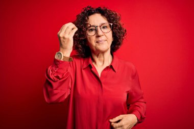 Orta yaşlı güzel kıvırcık saçlı kadın kırmızı arka planda günlük gömlek ve gözlük takıyor. İtalyan jestleri yapıyor.