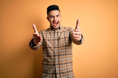 Sarı arka plan üzerinde duran rahat tişörtlü genç yakışıklı adam neşeli gülümseyerek yardım ve kabullenme sunuyor..