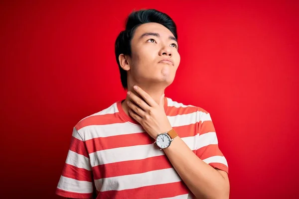 Young handsome chinese man wearing casual striped t-shirt standing over red background Touching painful neck, sore throat for flu, clod and infection