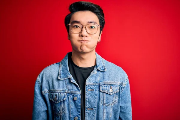 Young handsome chinese man wearing denim jacket and glasses over red background puffing cheeks with funny face. Mouth inflated with air, crazy expression.