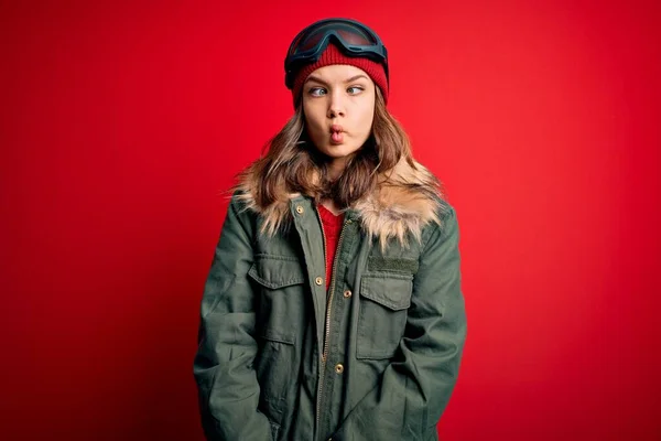年轻的金发女郎戴着滑雪眼镜 穿着冬衣在红色背景下滑雪 用嘴唇 疯狂和滑稽的姿势做着鱼脸 有趣的表达方式 — 图库照片
