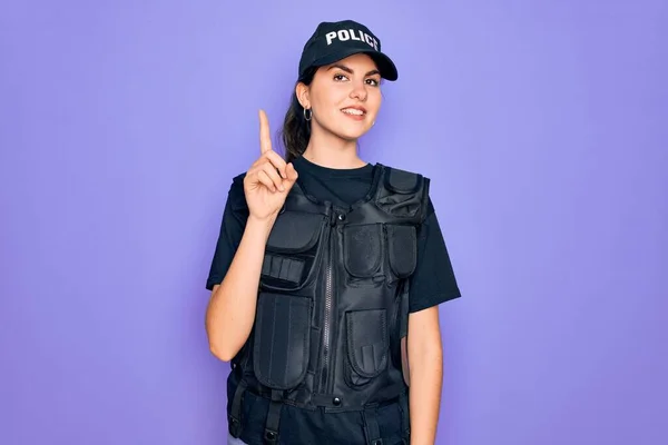 Young police woman wearing security bulletproof vest uniform over purple background pointing finger up with successful idea. Exited and happy. Number one.