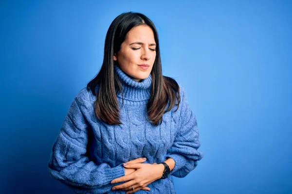 Young brunette woman with blue eyes wearing casual turtleneck sweater with hand on stomach because indigestion, painful illness feeling unwell. Ache concept.