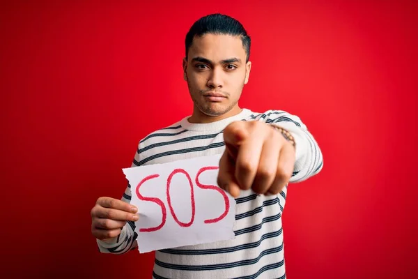 Young brazilian man with problem holding banner with sos message over red background pointing with finger to the camera and to you, hand sign, positive and confident gesture from the front