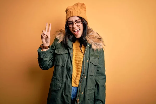 Young brunette woman wearing glasses and winter coat with hat over yellow isolated background smiling with happy face winking at the camera doing victory sign. Number two.