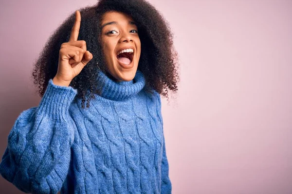 ピンクの背景の上に冬のセーターを着てアフロの髪を持つ若い美しいアフリカ系アメリカ人女性は 成功したアイデアで指を指しています 興奮し 第一番 — ストック写真
